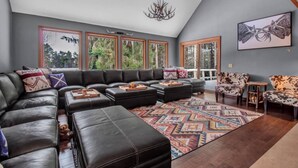 A spacious living room with a sectional sofa, ottomans, a patterned rug, and a large window with a view of trees.
