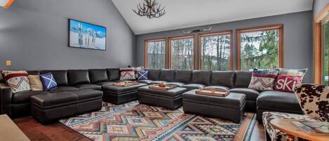 Un salon spacieux, doté d’un grand canapé d’angle, d’un tapis à motifs et offrant une vue sur l’extérieur grâce à de grandes fenêtres.