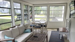 A sunroom with a white sofa, wooden coffee table, and chairs. Large windows offer views of the outside. - Meredith Bay - Brand New 1st Floor Apartment In Private Home (Meredith)