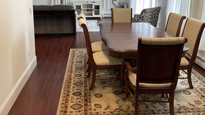 A dining area with a large wooden table, six chairs, a patterned rug, and a television on the wall. - Meredith Bay - Brand New 1st Floor Apartment In Private Home (Meredith)