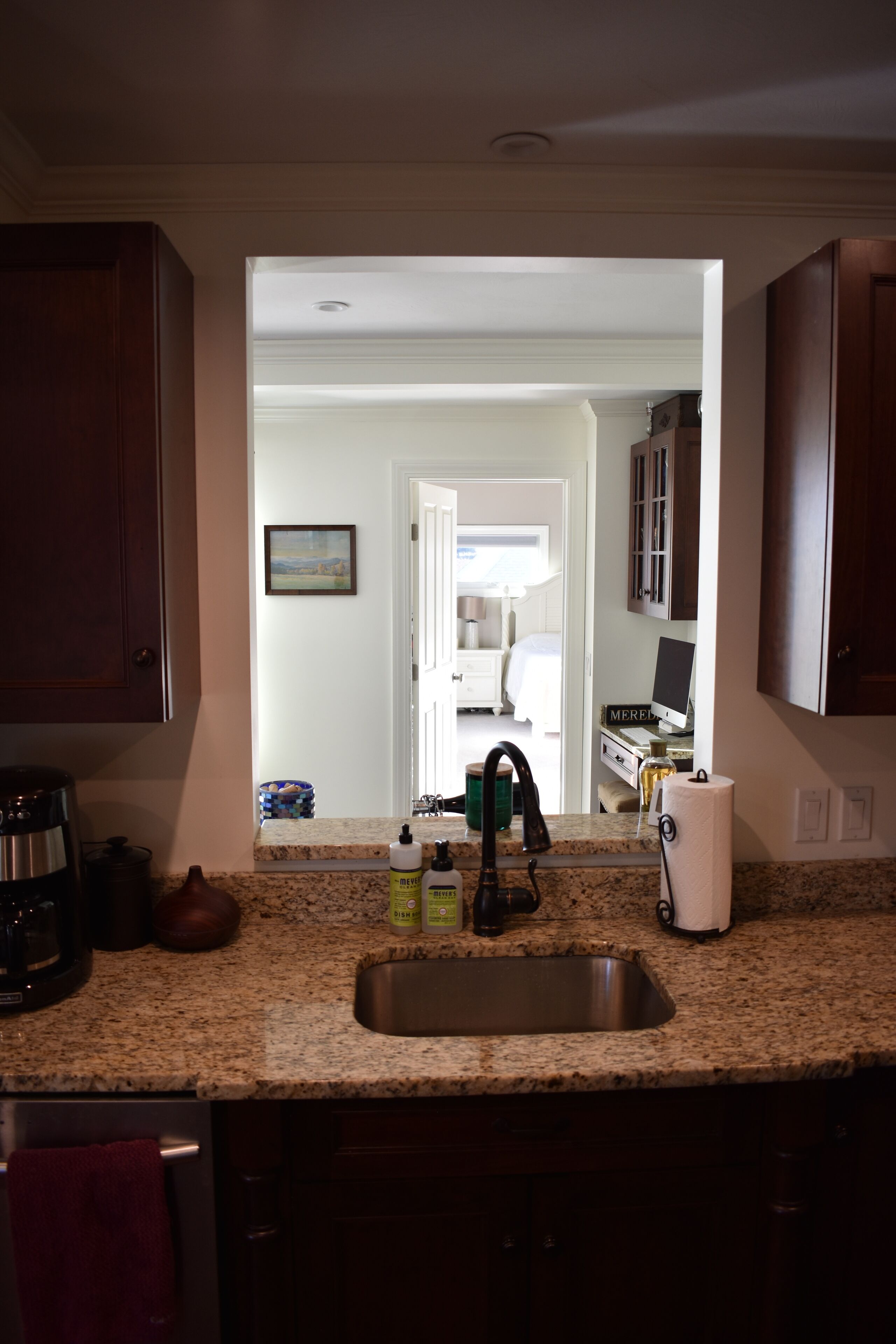 A kitchen with granite countertops, a stainless steel sink, and wooden cabinets. There is a coffee maker, a kettle, and a toaster on the counter. A bottle of hand soap and a paper towel dispenser are also visible.