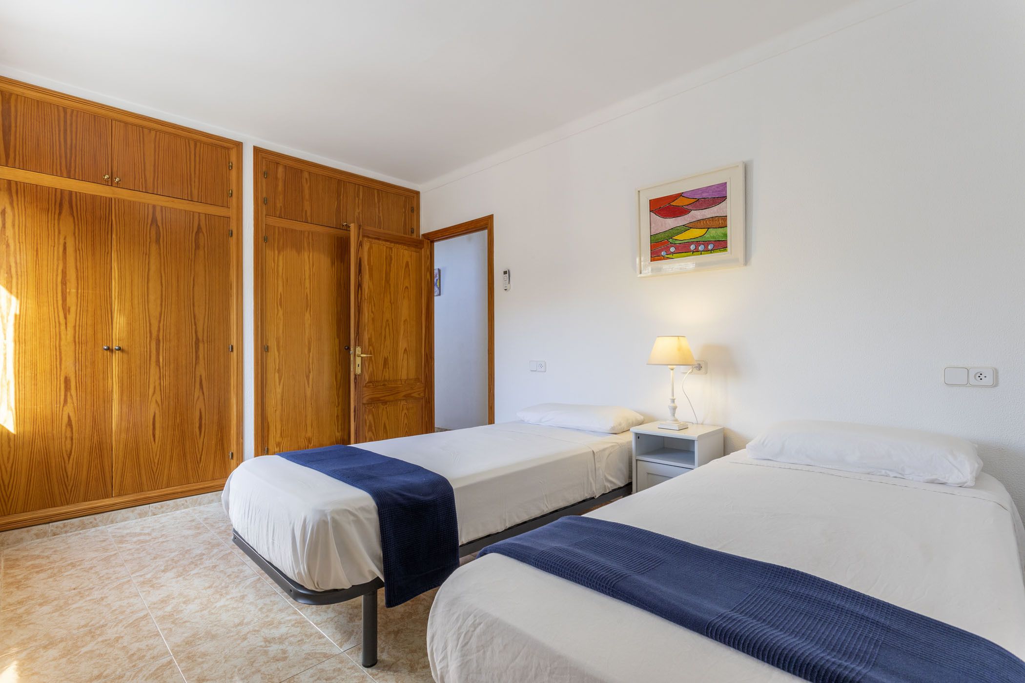 A room with two beds, wooden wardrobes, a nightstand with a lamp, and a colorful painting on the wall.