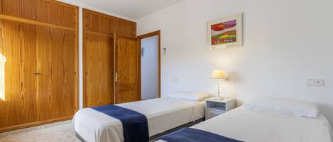 A room with two beds, wooden wardrobes, a nightstand with a lamp, and a colorful painting on the wall.