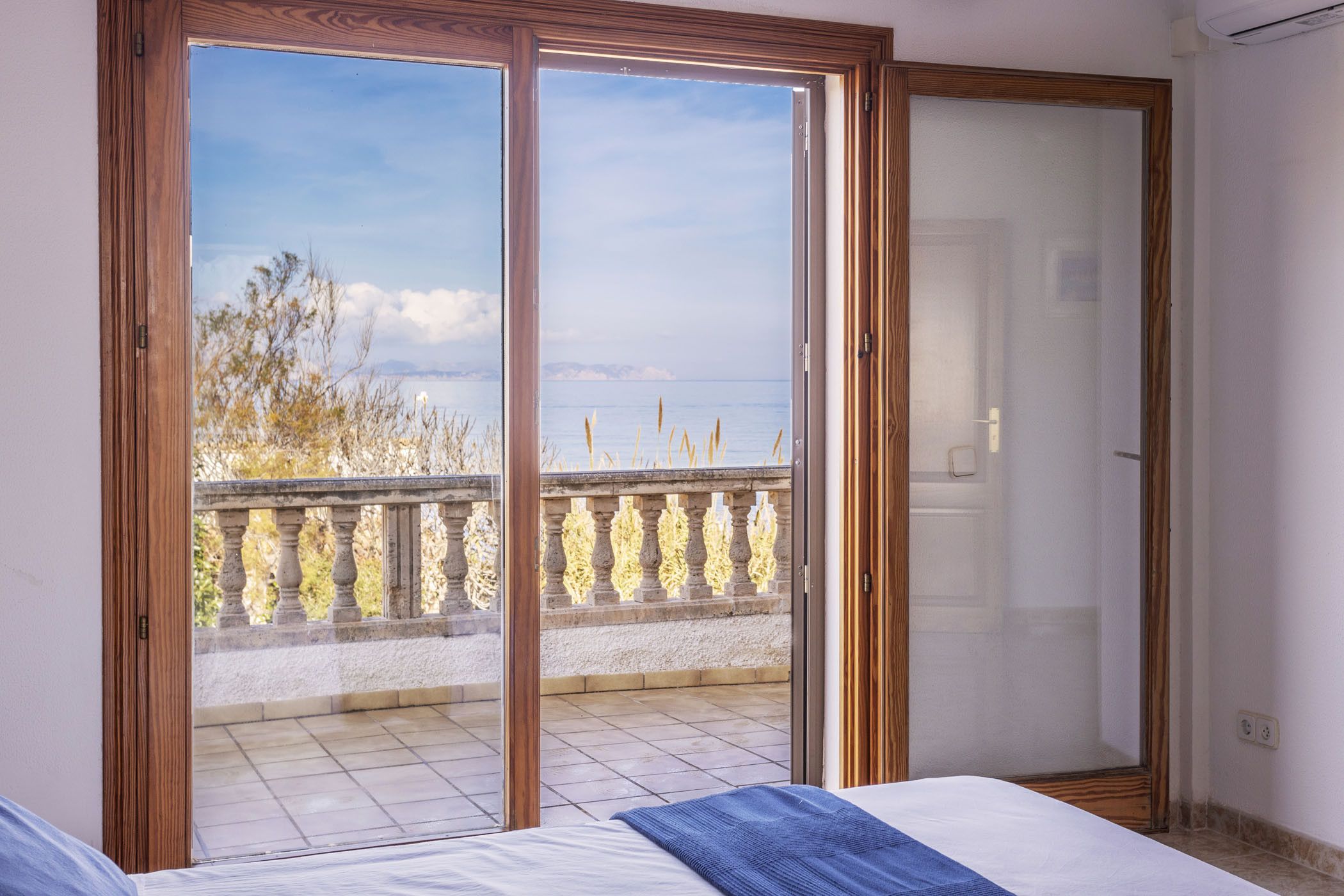A bedroom with a large sliding glass door leading to a balcony with a view of the sea and trees.