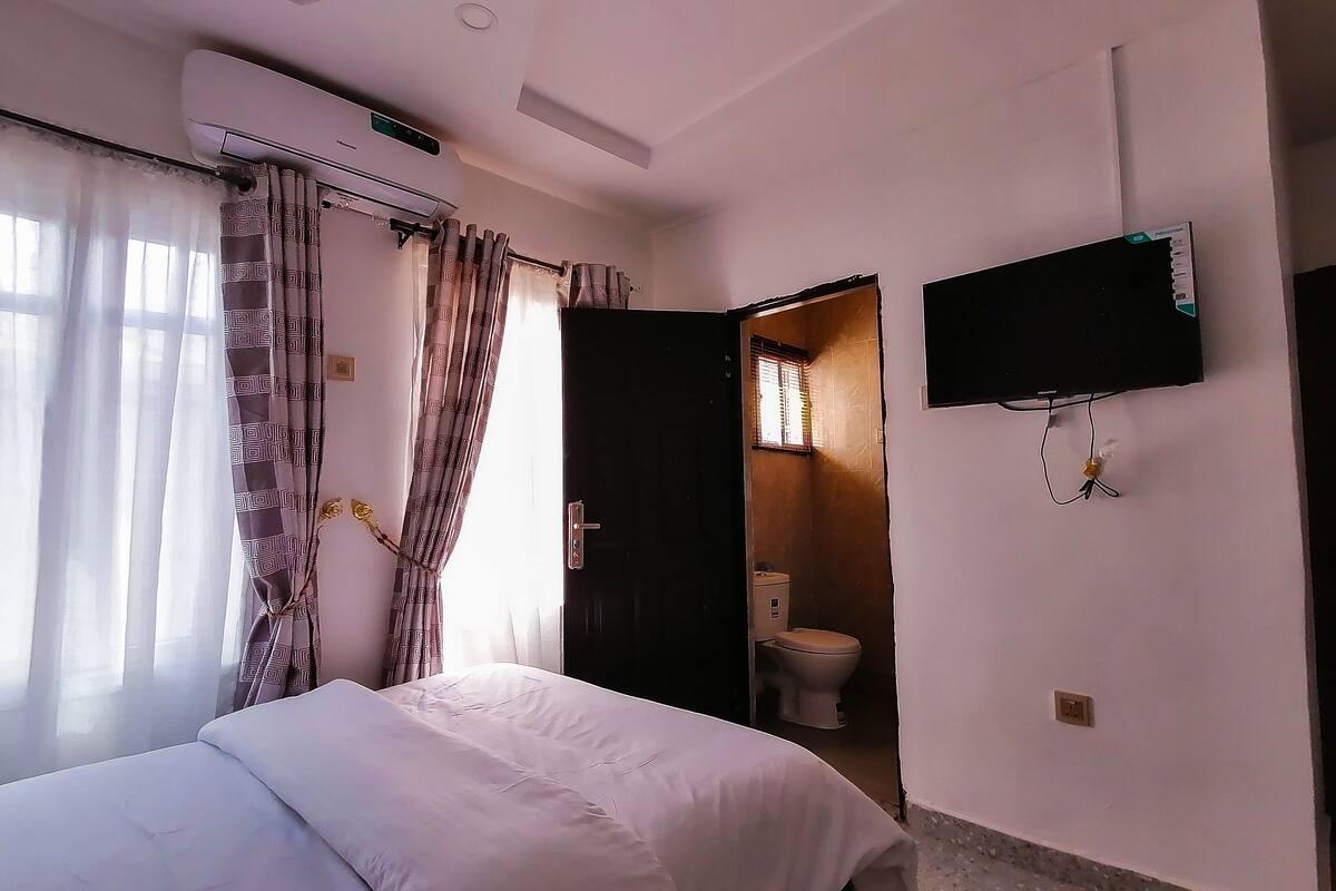 A hotel room with a bed, an air conditioning unit, a television mounted on the wall, and a bathroom visible through an open door.