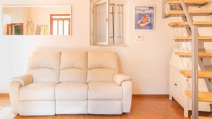 A living room with a beige sofa, a wooden staircase, and a window with a poster. - Bright Finalborgo Apartment - BBQ, Bike Storage, Garden (Finale Ligure)