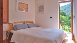 A bedroom with a wooden bed, a small bedside table, and a view of a mountainous landscape through an open door. - Bright Finalborgo Apartment - BBQ, Bike Storage, Garden (Finale Ligure)