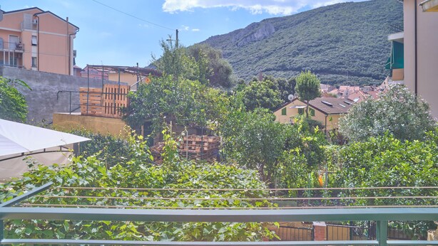 A balcony with a metal railing, a white awning, and a view of a hillside with buildings and greenery. - Bright Finalborgo Apartment - BBQ, Bike Storage, Garden (Finale Ligure)