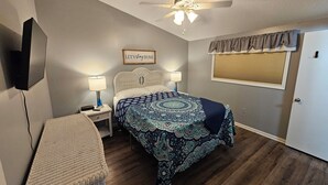 Une chambre avec un lit, deux lampes de chevet, un ventilateur de plafond, un téléviseur fixé au mur, une fenêtre avec des stores et un fauteuil en osier.