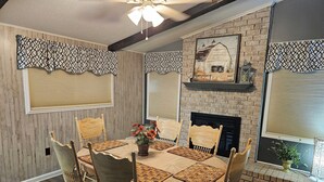 Une salle à manger avec une cheminée en briques, des lambrequins à motifs aux fenêtres et un ventilateur de plafond.