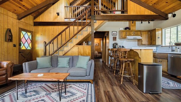 A cozy cabin interior with a wooden staircase, a living area with a sofa and coffee table, a kitchen with bar stools, and a large window. - Cabin that features modern upgrades near Snow & Play (Big Bear)