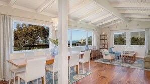 A spacious living area with a wooden floor, white furniture, and large windows offering a view of trees and hills. - Ocean & Mountain View Home - Walk to Trails, Beach, Family Activities (Montara)