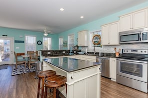 A kitchen with white cabinets, a central island, stainless steel appliances, and a view of the outside. - Dawn By The Bay-Designer Decor with Great Ocean Views, Sleeps 12 (Surf City)