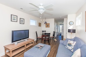 A living room with a flat-screen TV, a ceiling fan, a dining area with chairs, and a sofa with decorative pillows. - Dawn By The Bay-Designer Decor with Great Ocean Views, Sleeps 12 (Surf City)