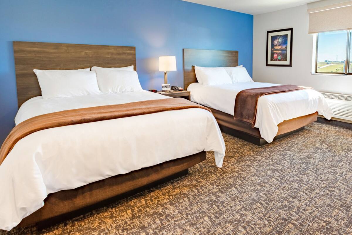 A hotel room with two beds, a nightstand with a lamp, a framed picture on the wall, and a window with a view.