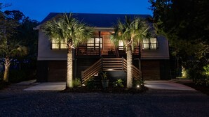 A modern two-story house with a wooden staircase, palm trees, and a well-lit entrance. - Pool, Hot Tub & Perfect Fall Beach Days (Isle of Palms)