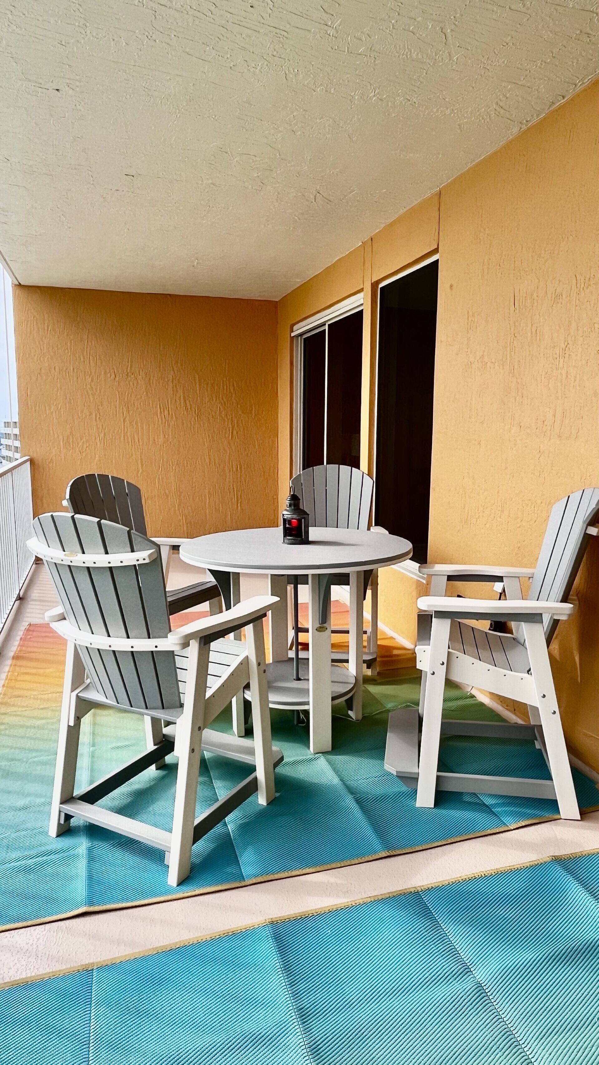 Un petit patio extérieur avec une table ronde et quatre chaises Adirondack, une lanterne et une vue sur un bâtiment à travers une fenêtre.