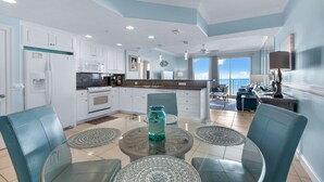 A modern kitchen with white cabinets, a central island, and a dining area with teal chairs and woven placemats.