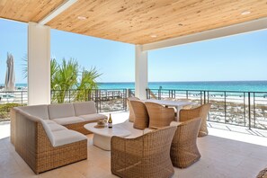 A covered outdoor seating area with wicker furniture, a white table, and a view of the beach.