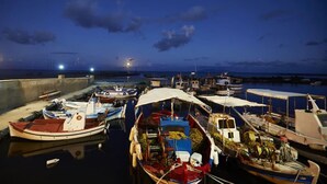 A marina at night with several boats docked, a concrete wall, and a calm sea under a dark sky with a visible moon. - Rodothea on the Beach. (Monolithos)