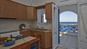 A compact kitchen with wooden cabinets, a stove, and a dining table with a bowl of apples. A window with curtains leads to a balcony with a view of the sea. - Rodothea on the Beach. (Monolithos)