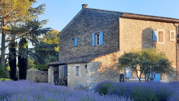 A stone building with a sloped roof, a small porch with outdoor seating, and a garden with lavender plants.