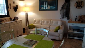 A cozy living room with a white sofa, a dining table with green placemats, a lamp, and a framed picture on the wall.