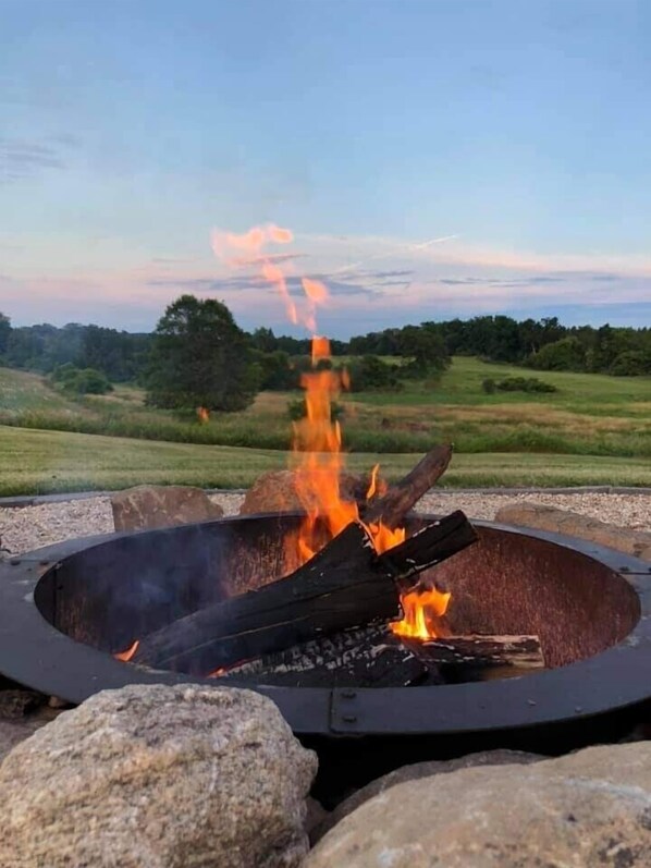A circular fire pit with burning logs and flames, surrounded by rocks, set in an outdoor area with a grassy field and trees in the background. - Restored Farmhouse Close to WI Dells (Oxford)