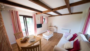 A cozy living room with a wooden dining table set for breakfast, a sofa with cushions, and a television on the wall. - Luxury Country Cottage with Access to Swimming Pool (High Halden)