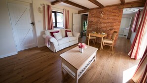 A cozy living room with a white sofa, a wooden coffee table, and a dining table with chairs. A brick wall and wooden beams are visible in the background. - Luxury Country Cottage with Access to Swimming Pool (High Halden)