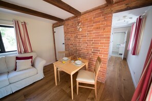 A cozy room with a brick wall, a wooden dining table with chairs, a white sofa, and a window with curtains. - Luxury Country Cottage with Access to Swimming Pool (High Halden)