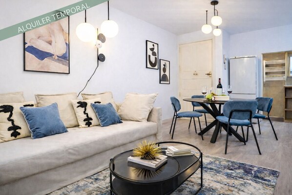 A modern living room with a sofa, coffee table, and dining area. There are framed artworks on the walls and a refrigerator in the background.