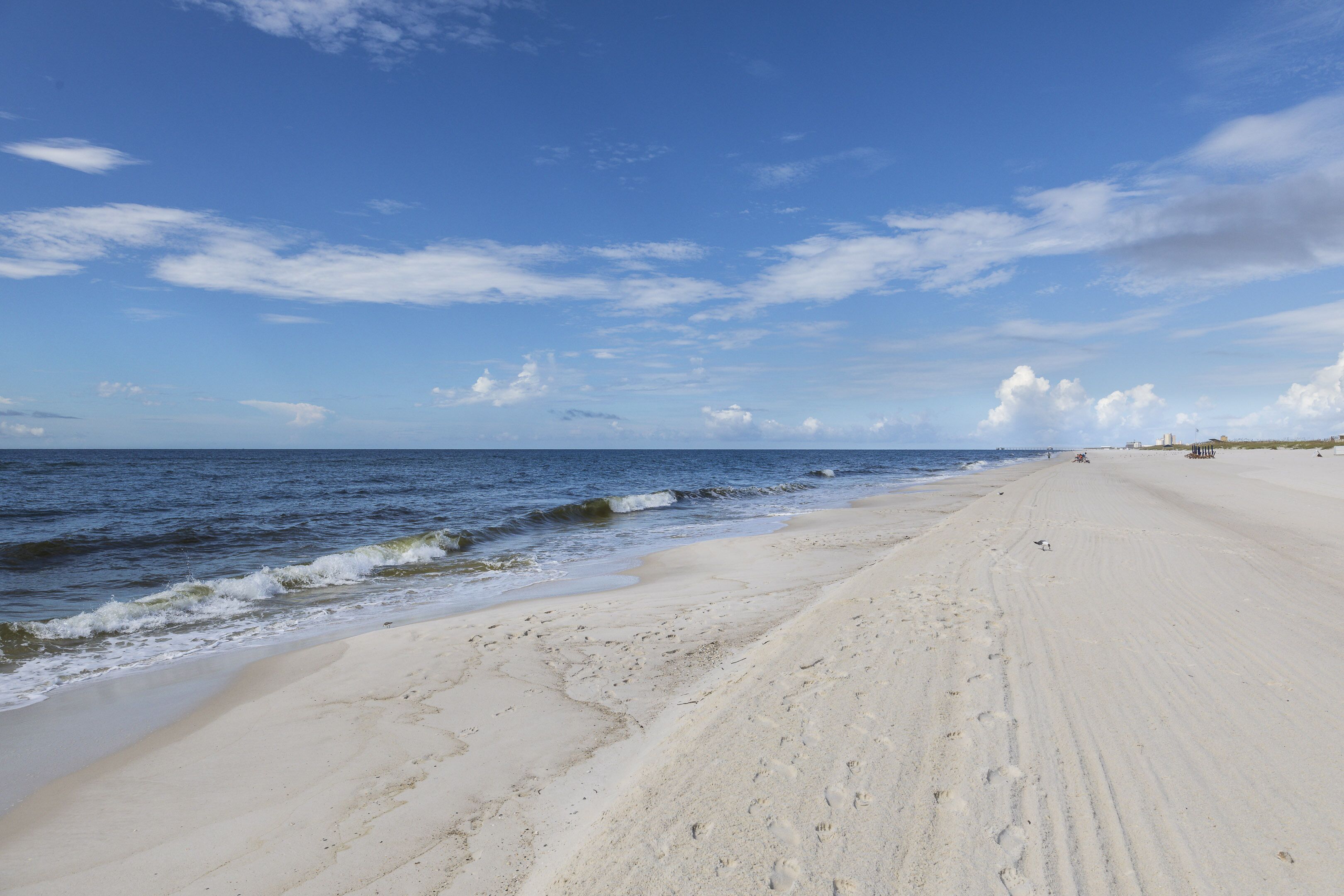 A wide sandy beach with gentle waves, a clear sky, and distant buildings.