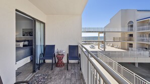 A balcony with a bed, two chairs, a small table, and a view of the beach and hotel buildings. - NEWLY RENOVATED CONDO- ON BEACH (Orange Beach)