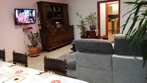 A living room with a grey sofa, a wooden cabinet, a television, and a dining table with a patterned tablecloth. - Gîte for 10 people (Prémanon)