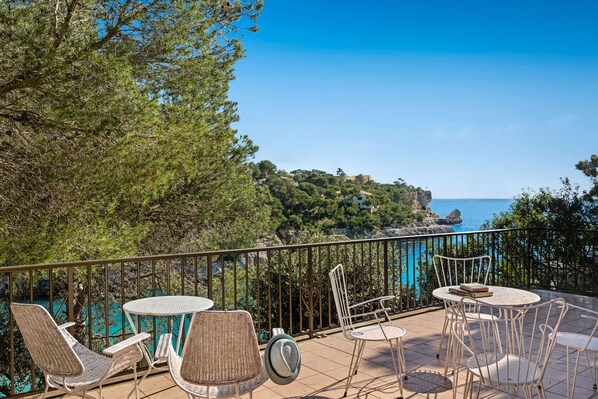 A terrace with wicker chairs and tables, overlooking a coastal landscape with trees and a view of the sea.
