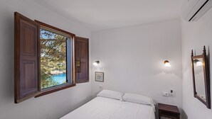A bedroom with a bed, a nightstand, a window with wooden shutters, and a wall-mounted light fixture. - Villa Sa Caleta (Cala Santanyí)