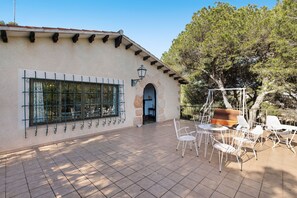 An outdoor patio area with tiled flooring, a seating arrangement with white chairs and tables, and a large tree providing shade.