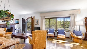 A spacious living room with a wooden floor, a large window with a view of greenery, and a variety of seating including a sofa, armchairs, and a dining table. - Villa Sa Caleta (Cala Santanyí)