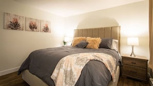 Un dormitorio con una cama grande, cabecera, mesita de noche con lámpara y tres cuadros de flores colgados en la pared.