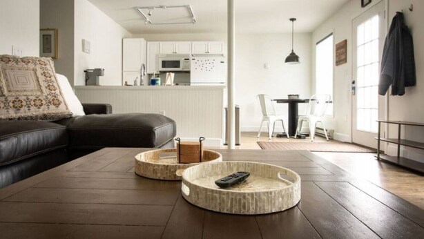 Una sala moderna con un sofá de cuero marrón oscuro, una mesa de centro redonda de madera y una cocina al fondo.