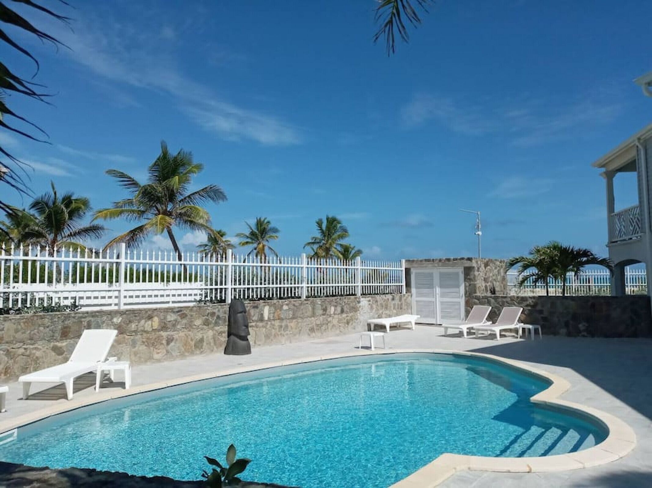 Une piscine d’un bleu éclatant, bordée de chaises longues blanches, de palmiers et d’une clôture blanche.