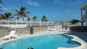 Una piscina azul cristalina con sillas de descanso blancas, palmeras y una valla blanca.