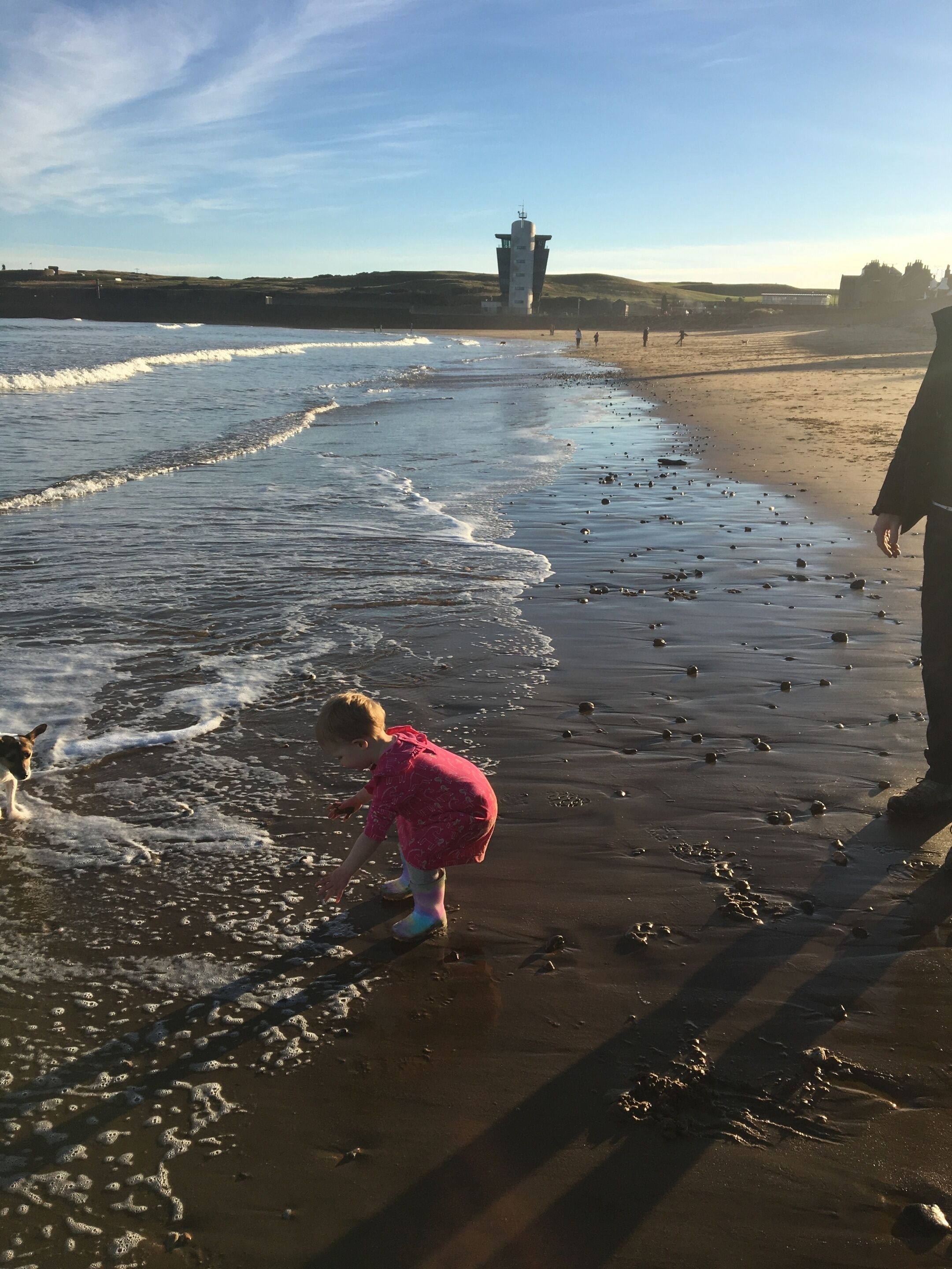 En strandscen med ett barn som leker i vattnet, en hund och en fyr i bakgrunden.
