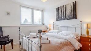 Un dormitorio con estructura metálica para la cama, dos camas, una ventana con persianas, una mesita de noche con lámpara, una silla y una obra de arte colgada en la pared.