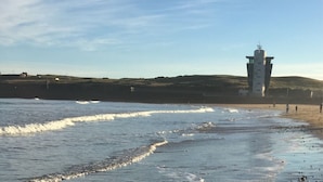 Paisaje costero con un faro, playa y colinas al fondo.