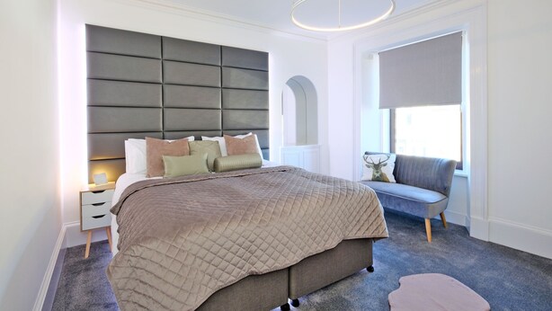 A bedroom with a large bed, a bedside table, a grey sofa, and a window with a view. - Ben Shea Smart Home (Keith)