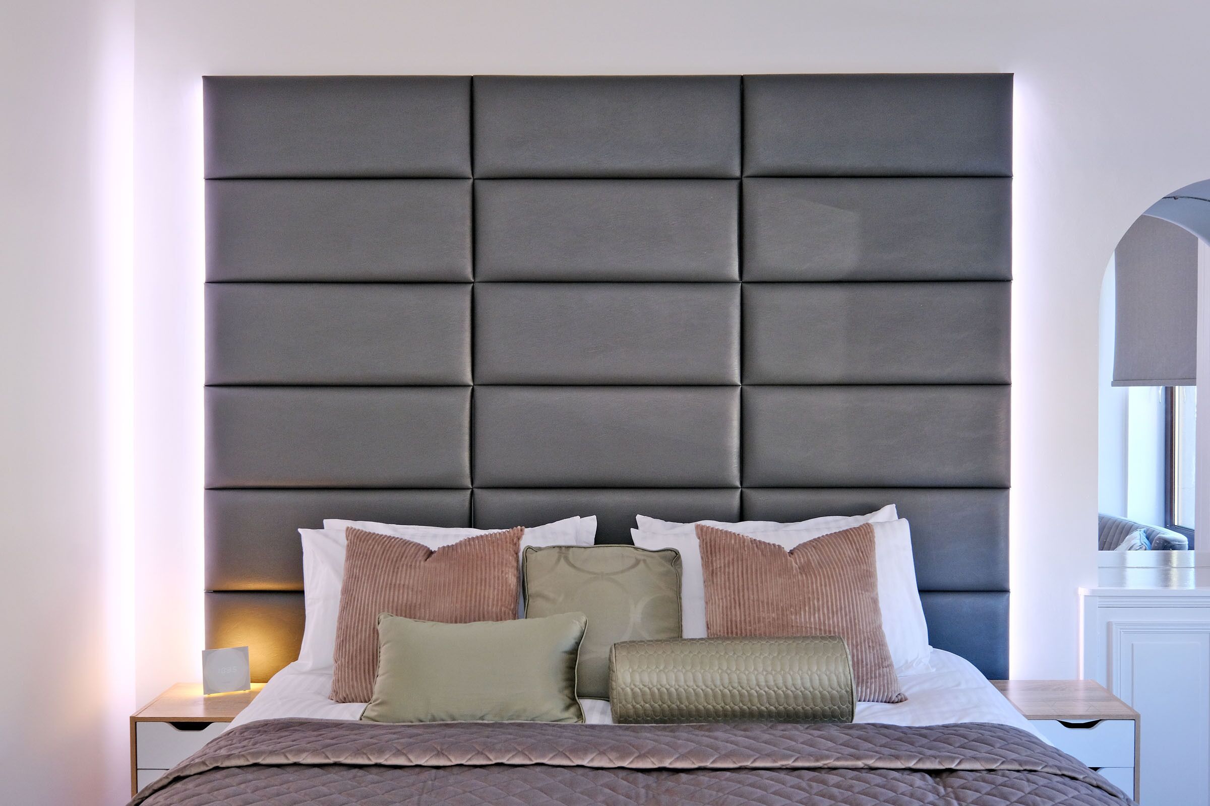Un lit bien fait, avec une tête de lit rembourrée, une table de chevet avec une lampe et un miroir au mur.