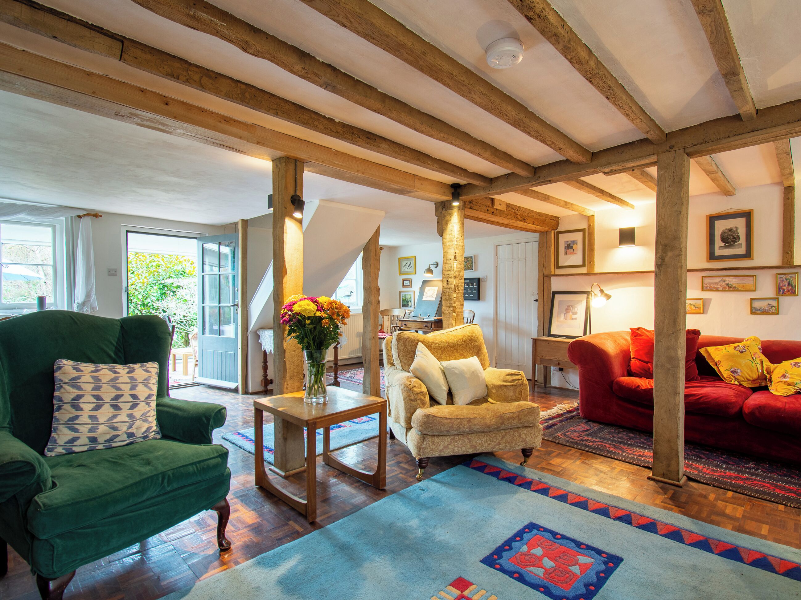 A room with a green armchair, a red sofa, wooden beams, a wooden table with a vase, and a patterned rug.