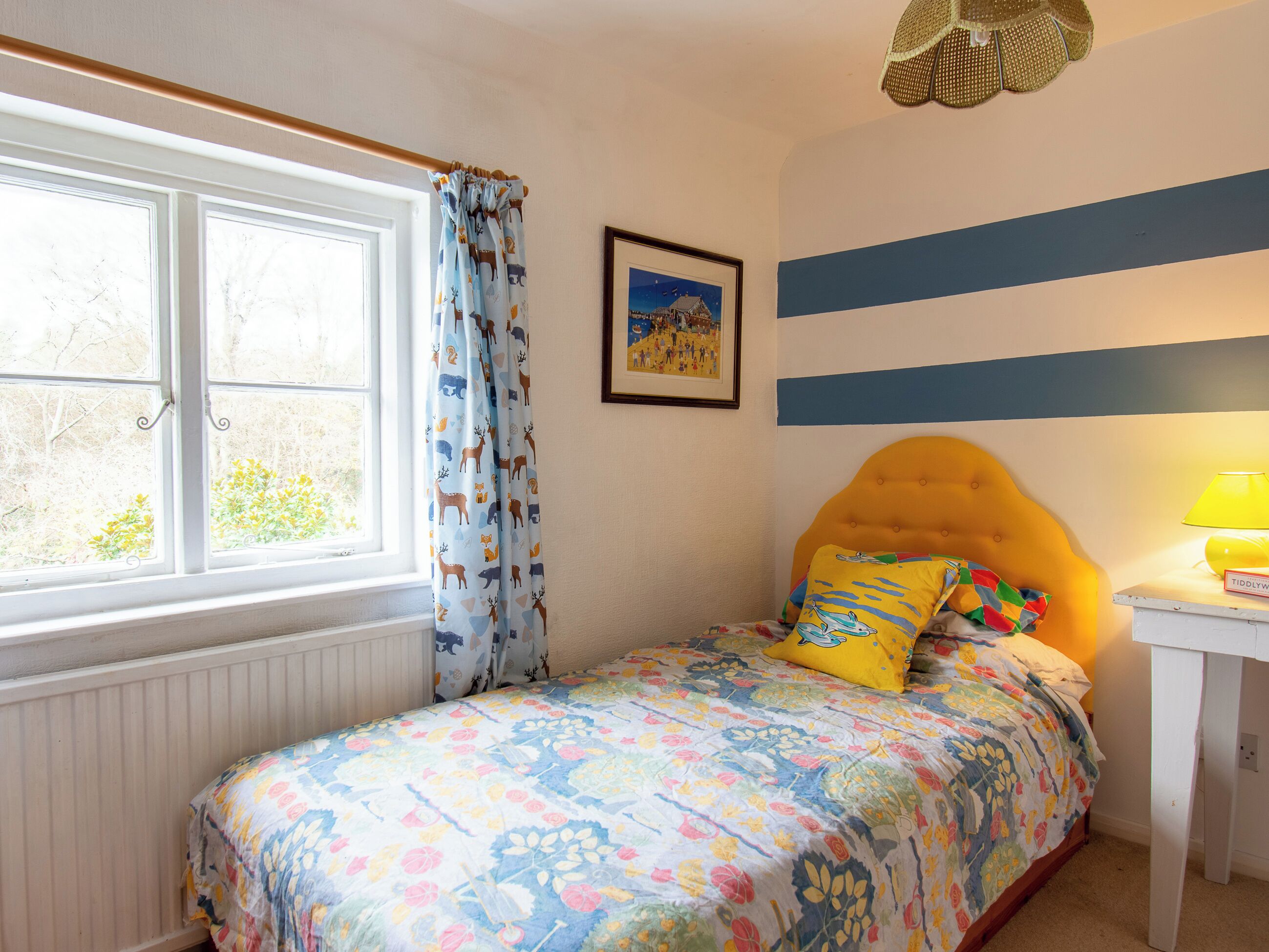 A bedroom with a bed, a window with curtains, a bedside table with a lamp, and a wall with horizontal stripes.